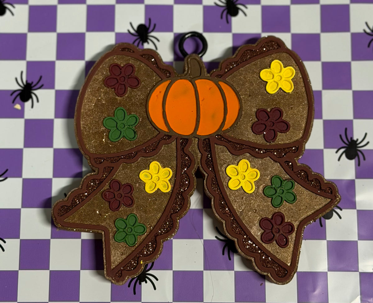 A decorative autumn-themed bow ornament with a glittery brown body, an orange pumpkin in the center, and embroidered-style flowers in yellow, green, and dark purple, displayed on a purple and white checkered background with black spider graphics.